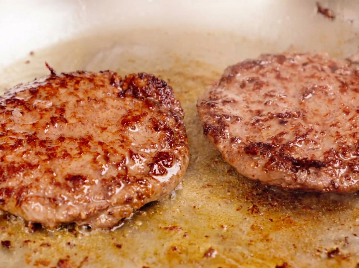 Zwei dicke Rindfleisch-Pattys braten in einer Pfanne, mit einer stark gebräunten Kruste und blubbernden Säften auf der Oberfläche.