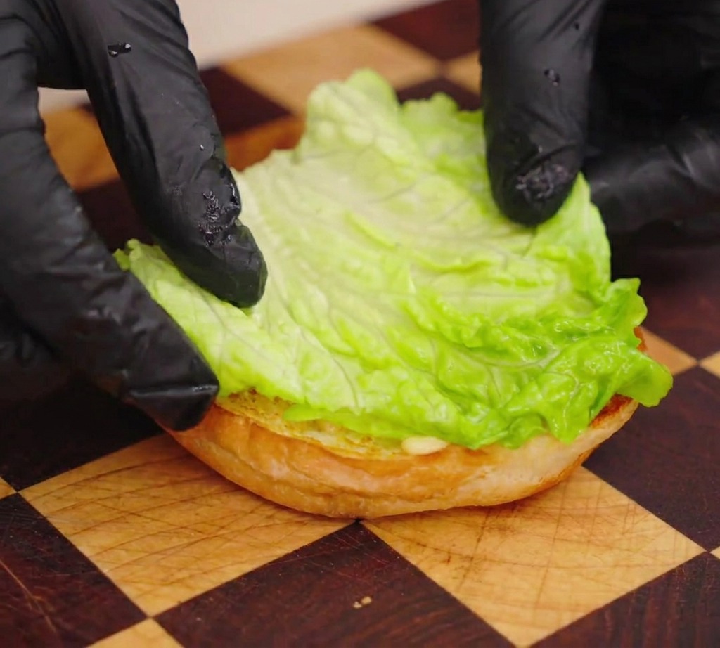 Eine Person mit schwarzen Handschuhen legt ein leuchtend grünes Salatblatt auf die untere Hälfte eines gerösteten Burgerbrötchens.