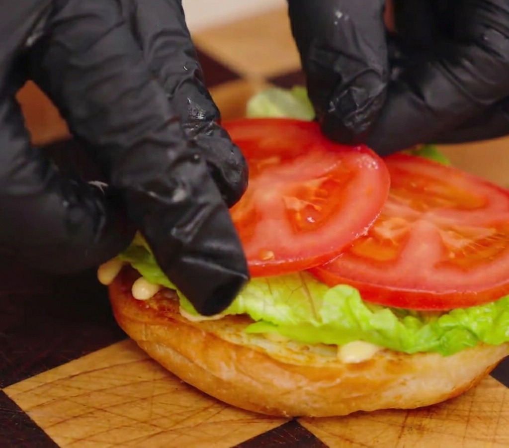 Nahaufnahme von bekehrten Händen, die zwei dicke, saftige rote Tomatenscheiben auf ein Salatblatt auf einem gerösteten Brötchen legen.