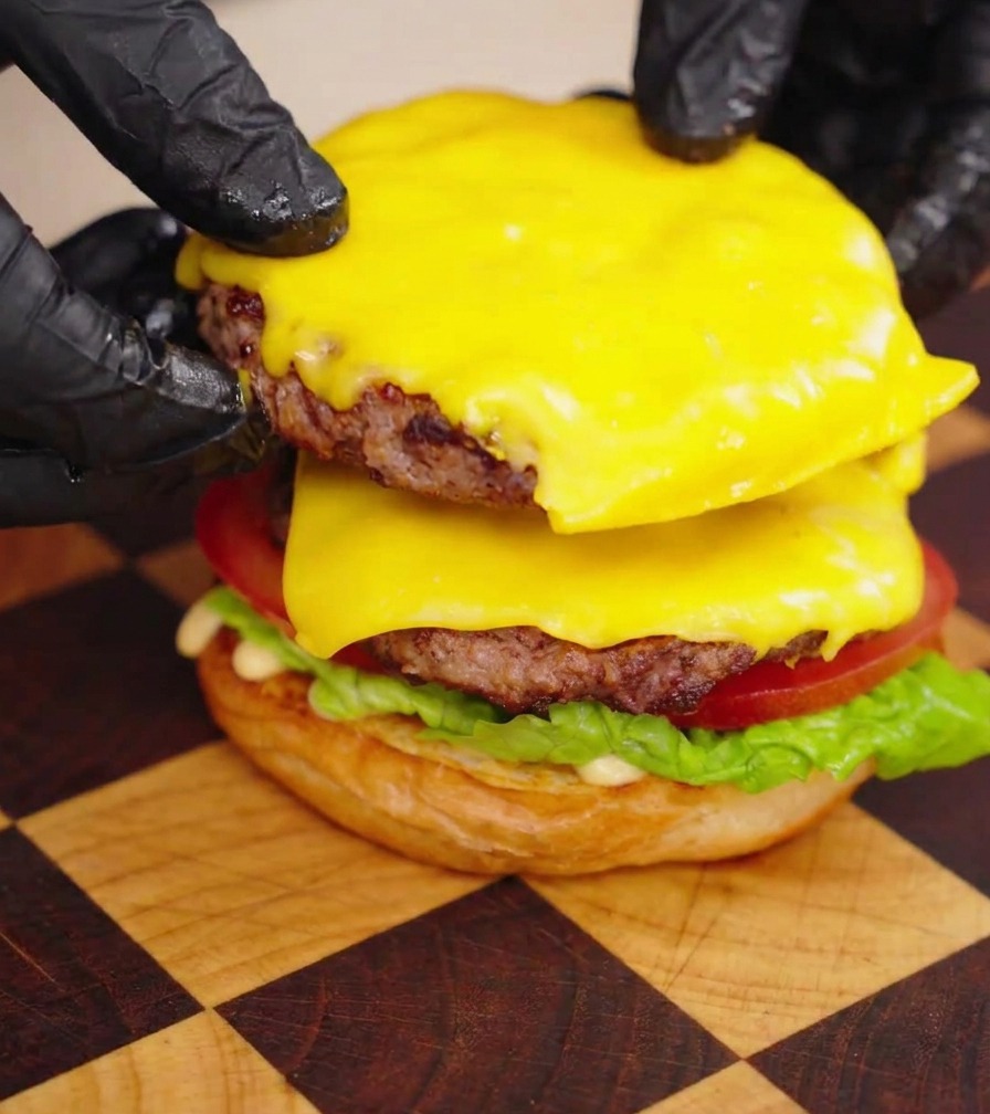 Hände in schwarzen Handschuhen stapeln zwei saftige, mit geschmolzenem Käse überzogene Wagyu-Pattys auf den Burger.
