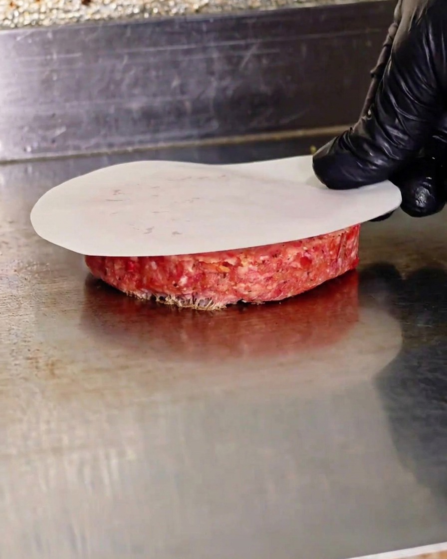 Eine behandschuhte Hand drückt ein rohes Rindfleisch-Patty mit einer flachen Presse über einem Stück Backpapier auf einer heißen Grillplatte platt.