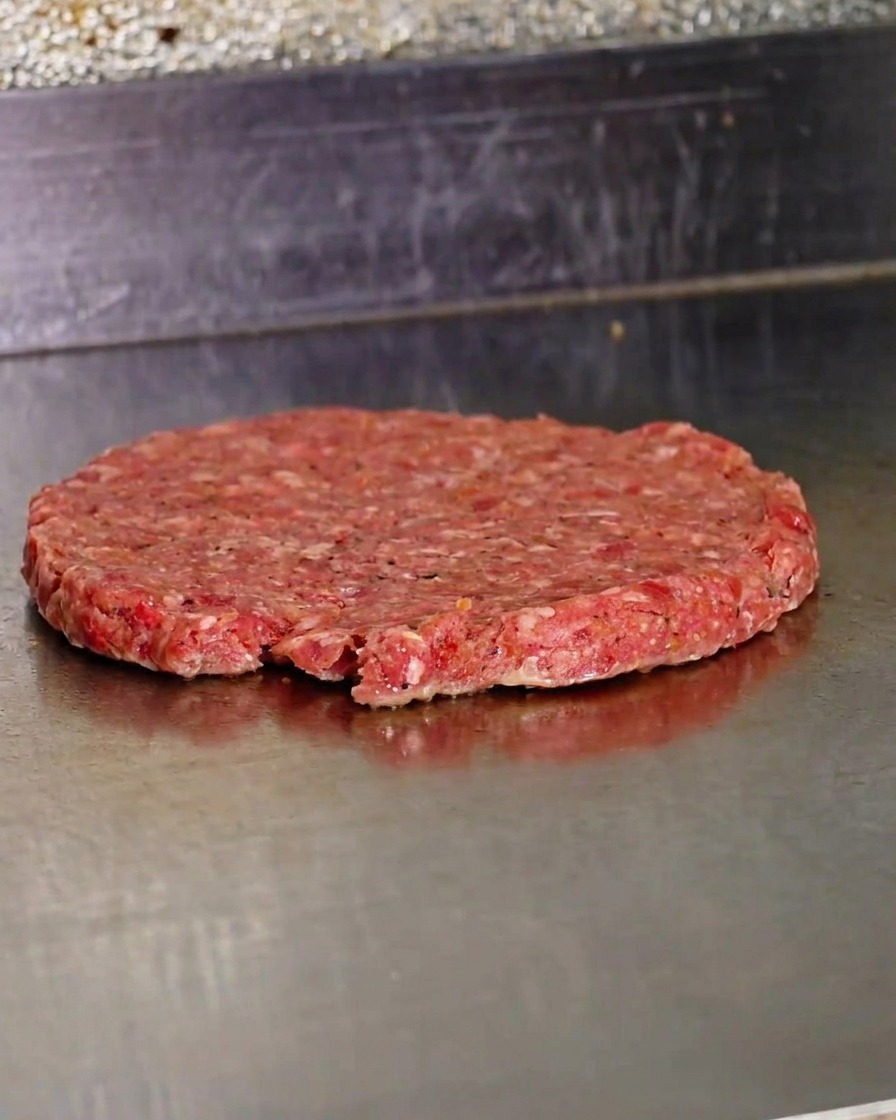 Ein frisch plattgedrücktes, rohes Rindfleisch-Patty brutzelt auf einer heißen Grillplatte.