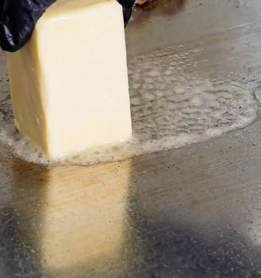 Eine Hand mit schwarzem Handschuh reibt ein rechteckiges Stück Butter auf eine heiße Grillplatte aus Metall, wodurch Bläschen aus schmelzendem Fett entstehen.