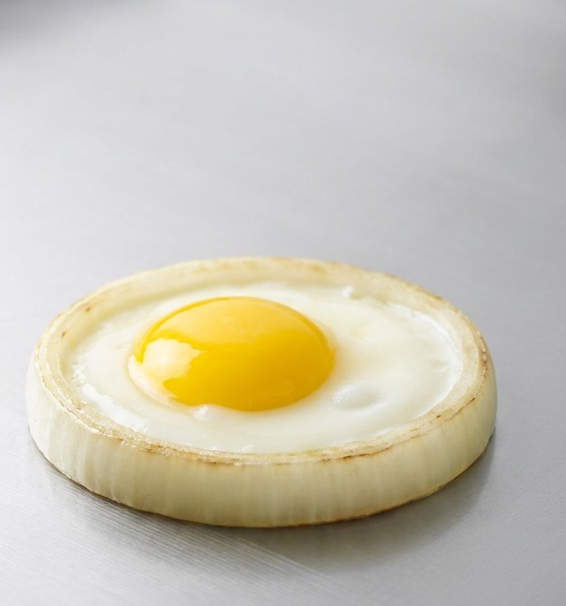 Ein perfekt kreisrundes Spiegelei mit leuchtend gelbem Eigelb brät in einem dicken Zwiebelring auf einer flachen Edelstahloberfläche.