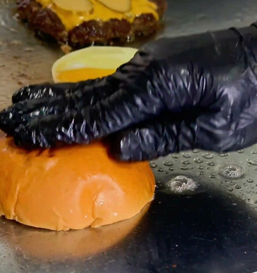 Eine behandschuhte Hand drückt ein Burger-Brötchen mit der Schnittfläche nach unten in eine Pfütze aus geschmolzener Butter auf einer Grillplatte neben einem bratenden Patty.