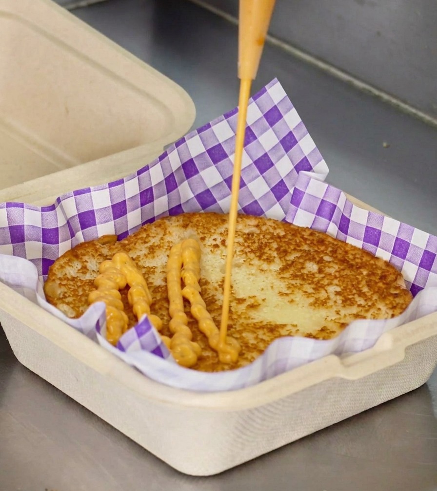 Ein Strahl cremiger, oranger Sauce wird aus einer Flasche auf die Mitte eines gerösteten Burger-Brötchens in einer lilafarben karierten Box-Einlage gedrückt.