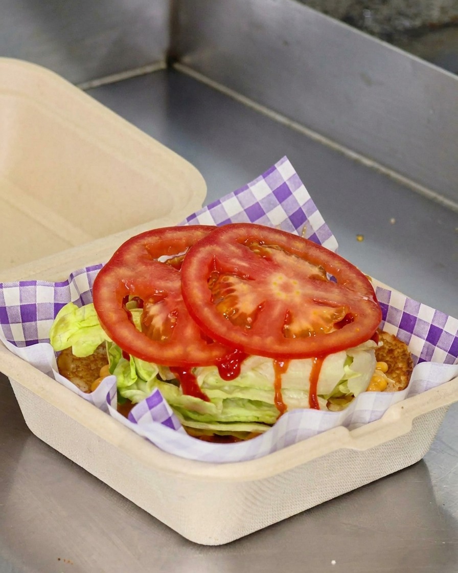 Zwei runde Scheiben frischer roter Tomate liegen auf zerkleinertem Salat in einem Takeaway-Behälter aus Papier.