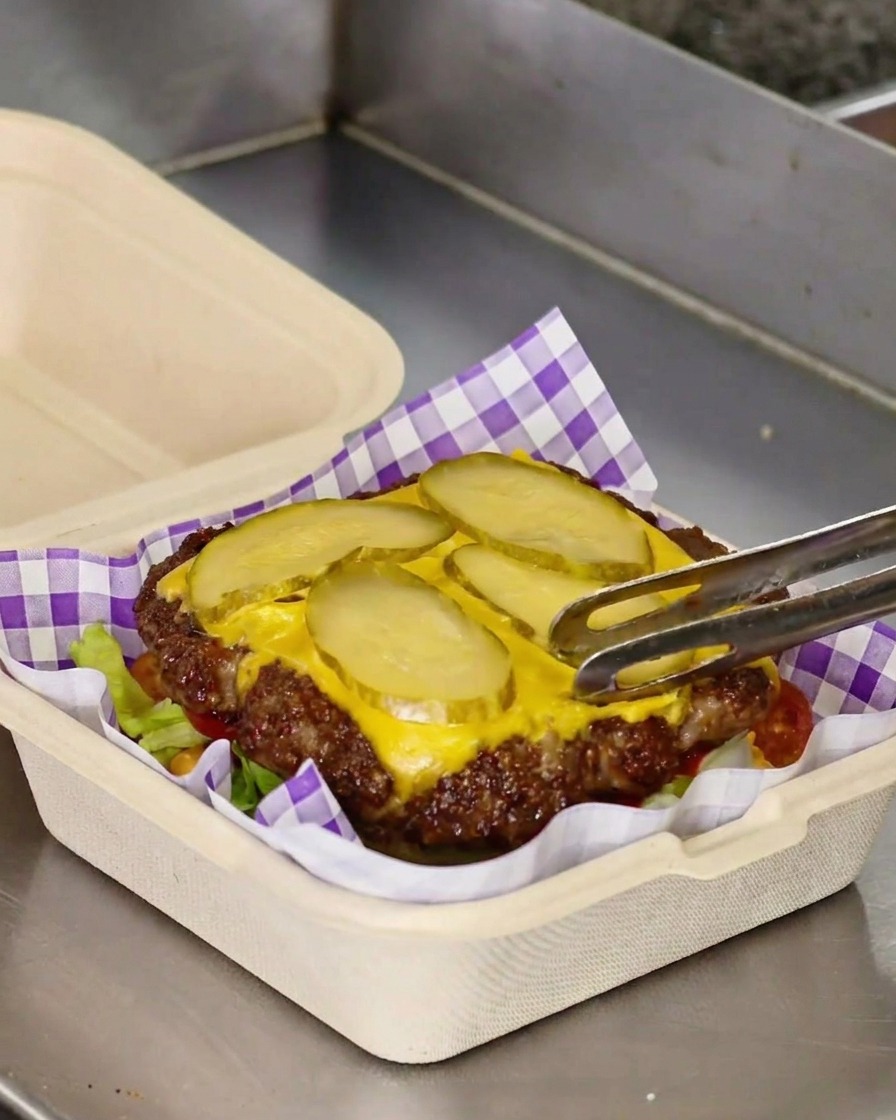Eine Zange überträgt ein gebratenes Smash-Burger-Patty, belegt mit geschmolzenem gelben Käse und Gurkenscheiben, auf ein Bett aus grünem Salat.