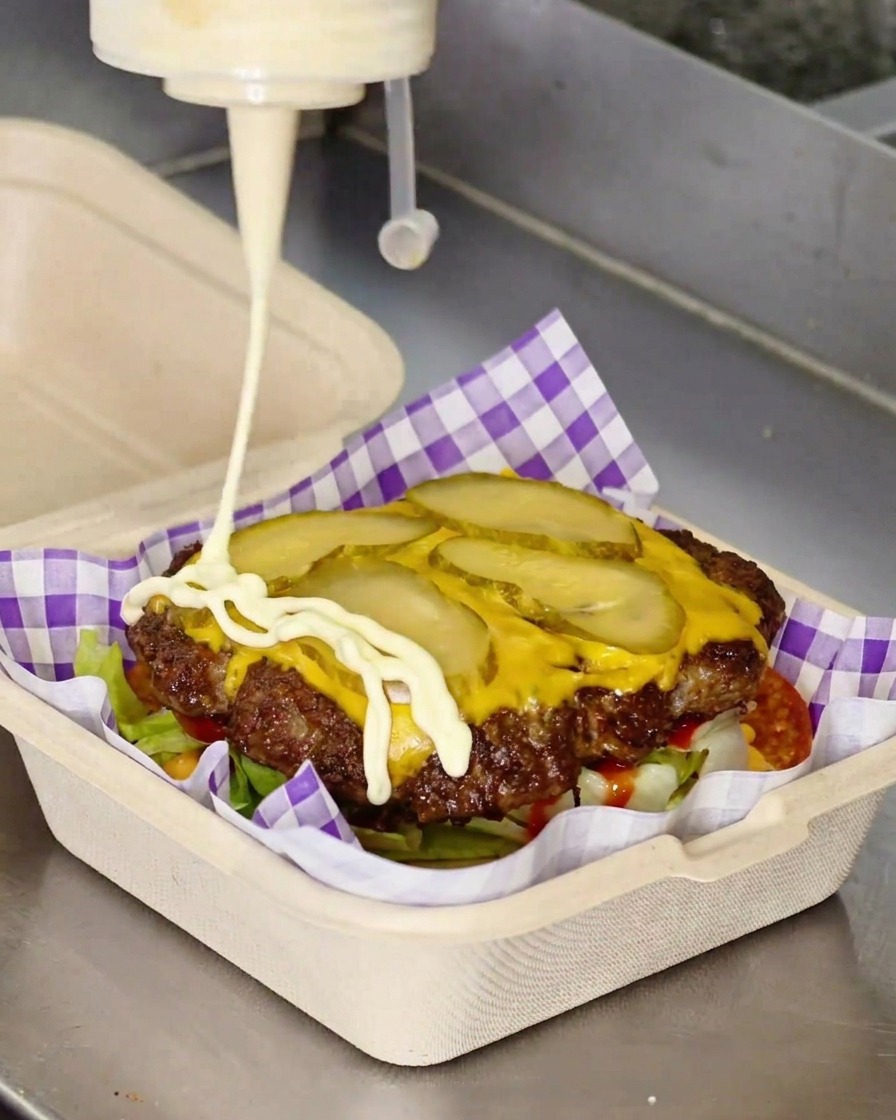 Weiße Mayonnaise wird in einem Zickzack-Muster aus einer Flasche über ein Cheeseburger-Patty gespritzt.
