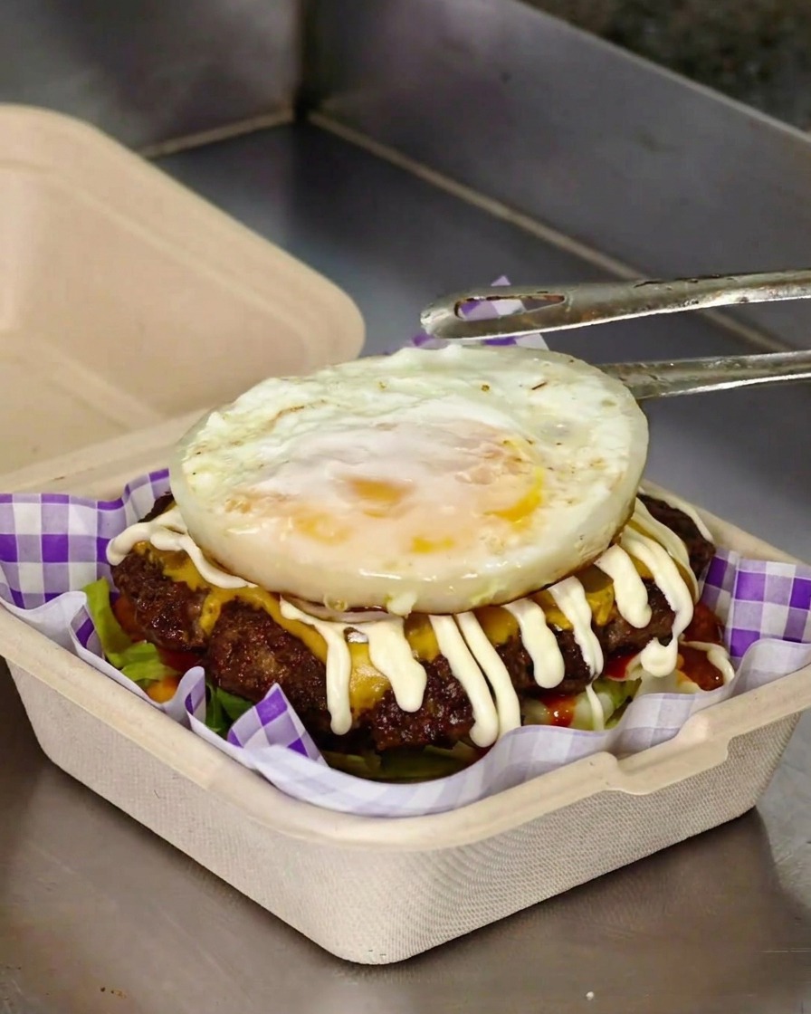 Eine Metallzange legt ein frisch gebratenes Spiegelei oben auf einen Burger, der mit plattgedrückten Fleisch-Pattys, geschmolzenem Käse und einem Zickzack aus Mayonnaise belegt ist.