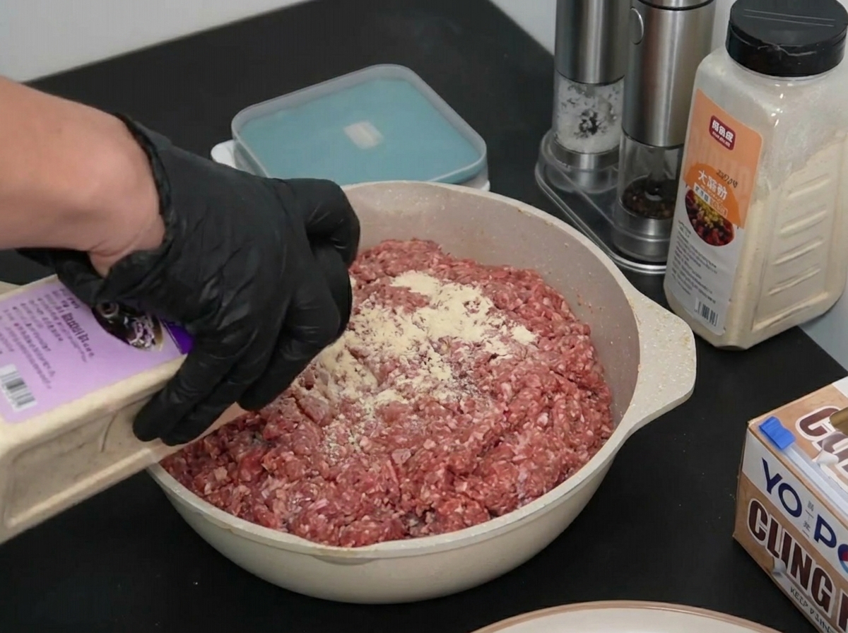 Eine behandschuhte Hand streut Zwiebelpulver aus einem Behälter über rohes Rinderhackfleisch in einer Form.