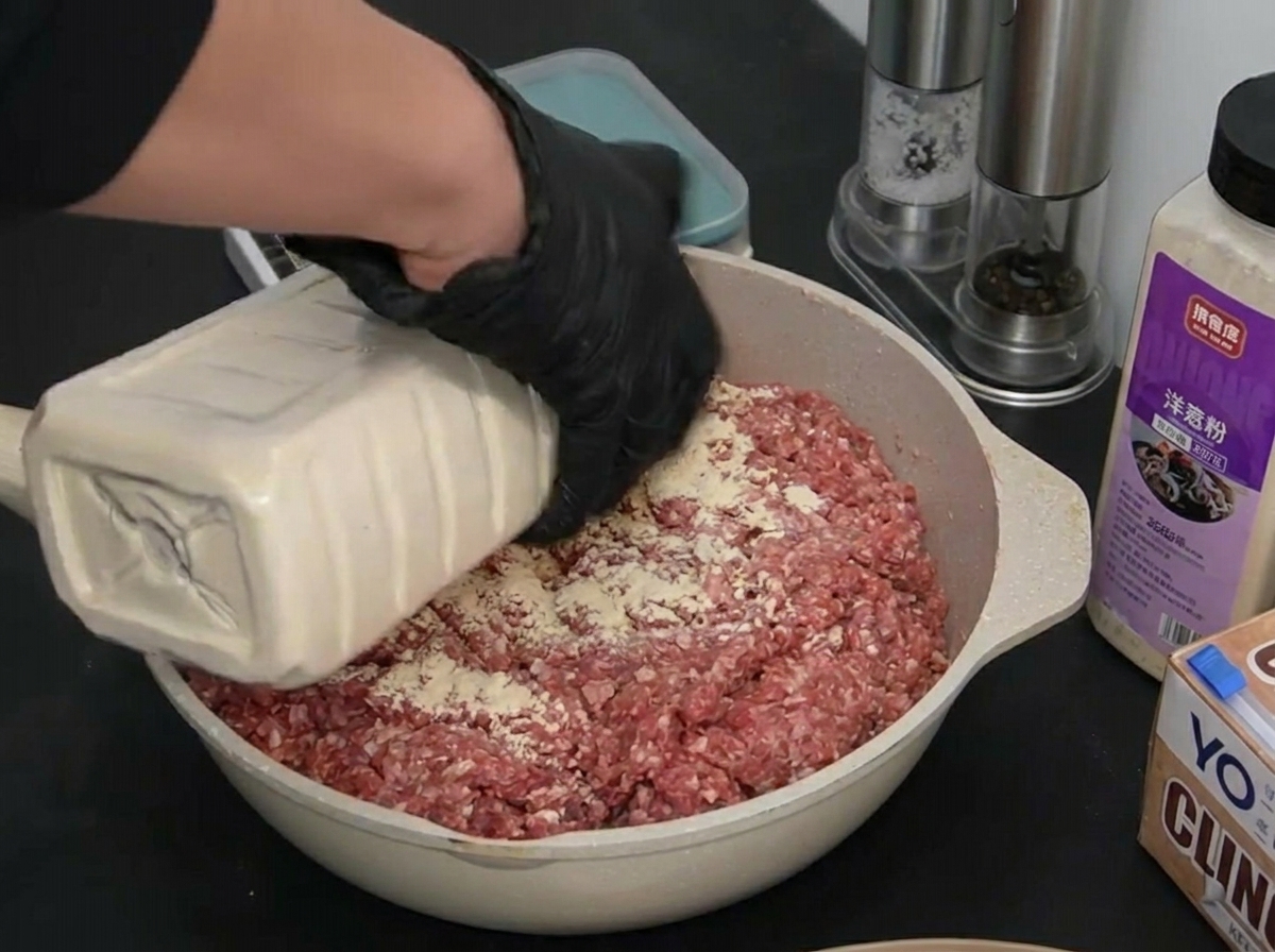 Eine Person mit schwarzen Handschuhen gießt weißes Knoblauchpulver über das flach ausgelegte Hackfleisch.