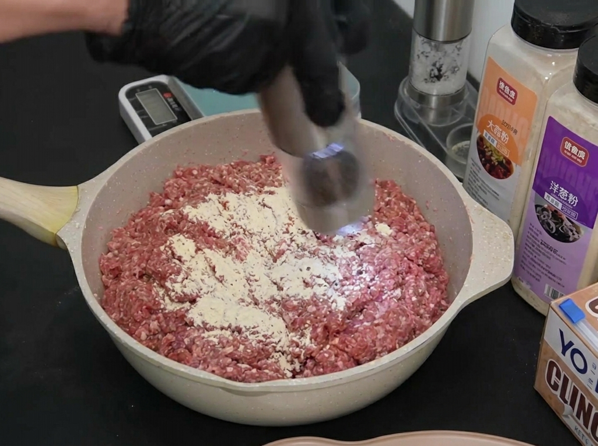 Eine behandschuhte Hand nutzt eine elektrische Mühle, um schwarzen Pfeffer über das Fleisch zu mahlen.