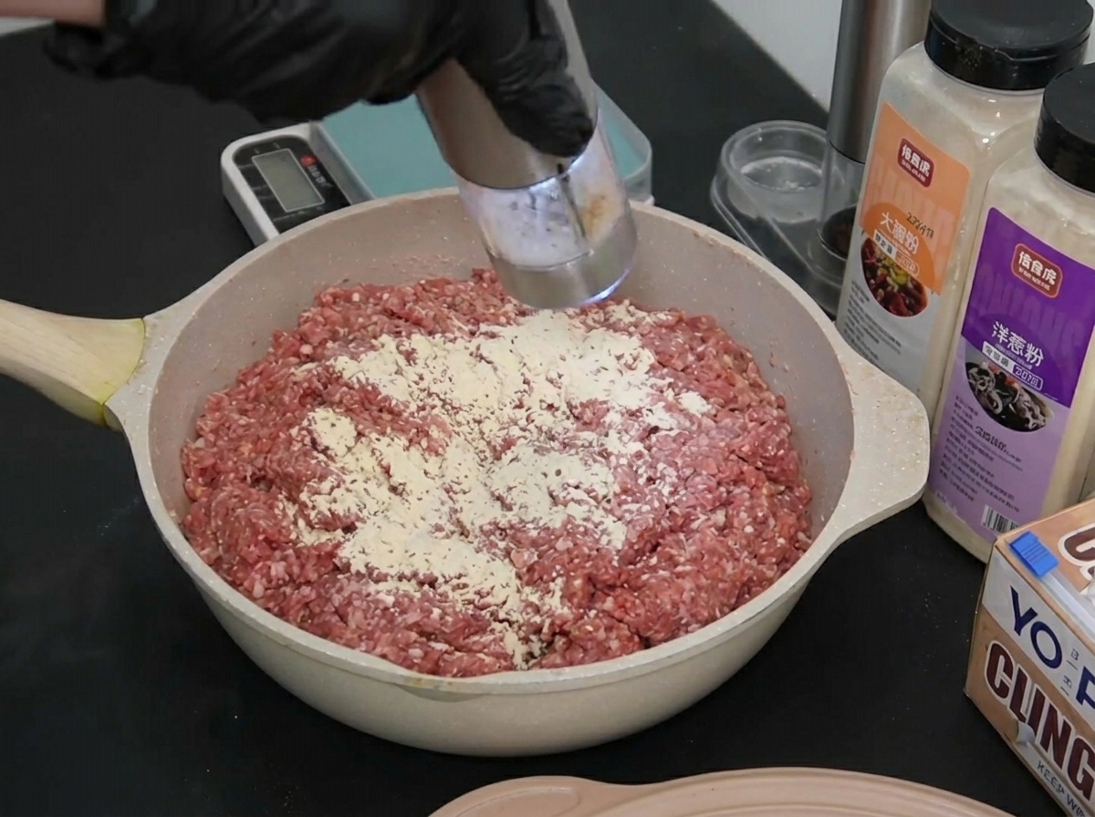 Eine behandschuhte Hand mahlt eine kleine Menge weißes Meersalz auf das gewürzte Hackfleisch.