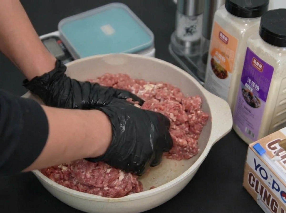 Zwei behandschuhte Hände kneten das gewürzte Hackfleisch in einer flachen Schale.