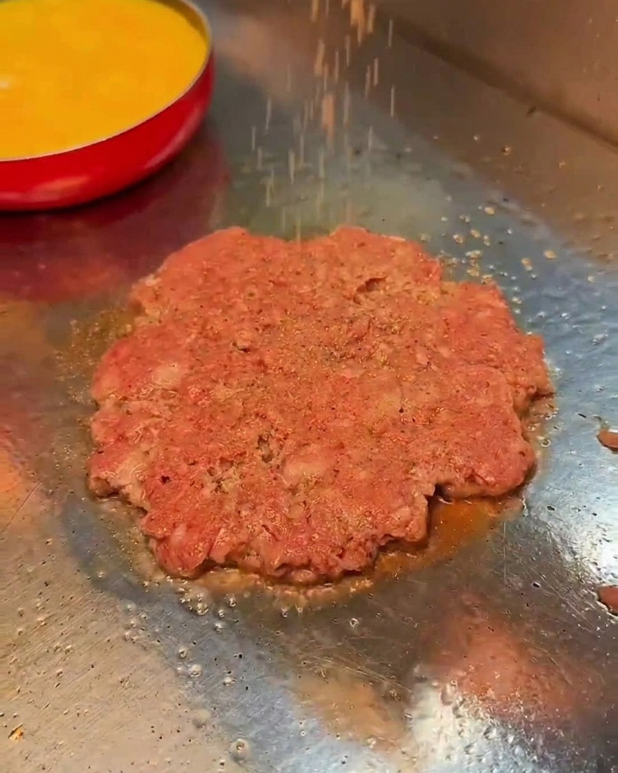 Salz und schwarzer Pfeffer werden über ein Fleisch-Patty auf dem Grill gestreut.