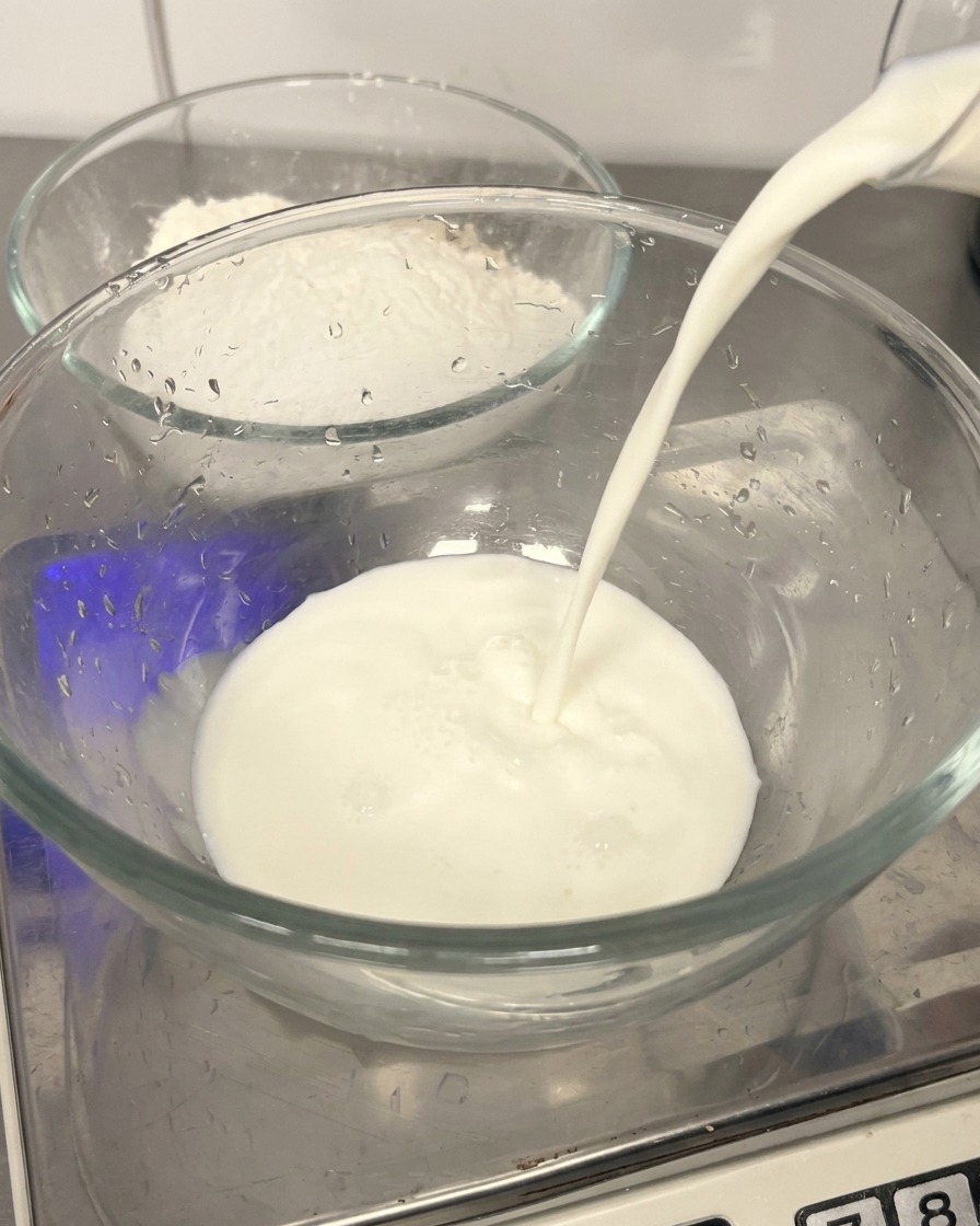 Frische Milch wird in eine Glasschüssel gegossen, im Hintergrund steht eine Schüssel mit Mehl.