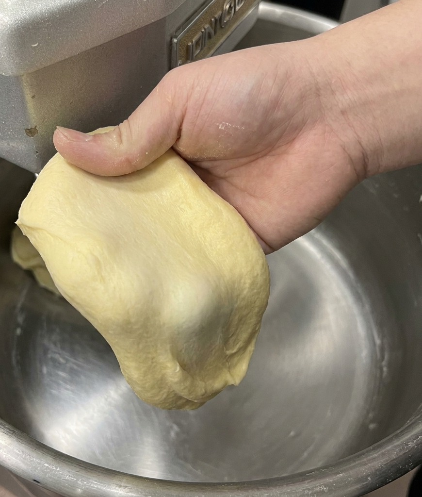 Eine Hand dehnt ein Stück hellgelben Brioche-Teig, um die dünne, starke Fensterscheiben-Membran zu zeigen.