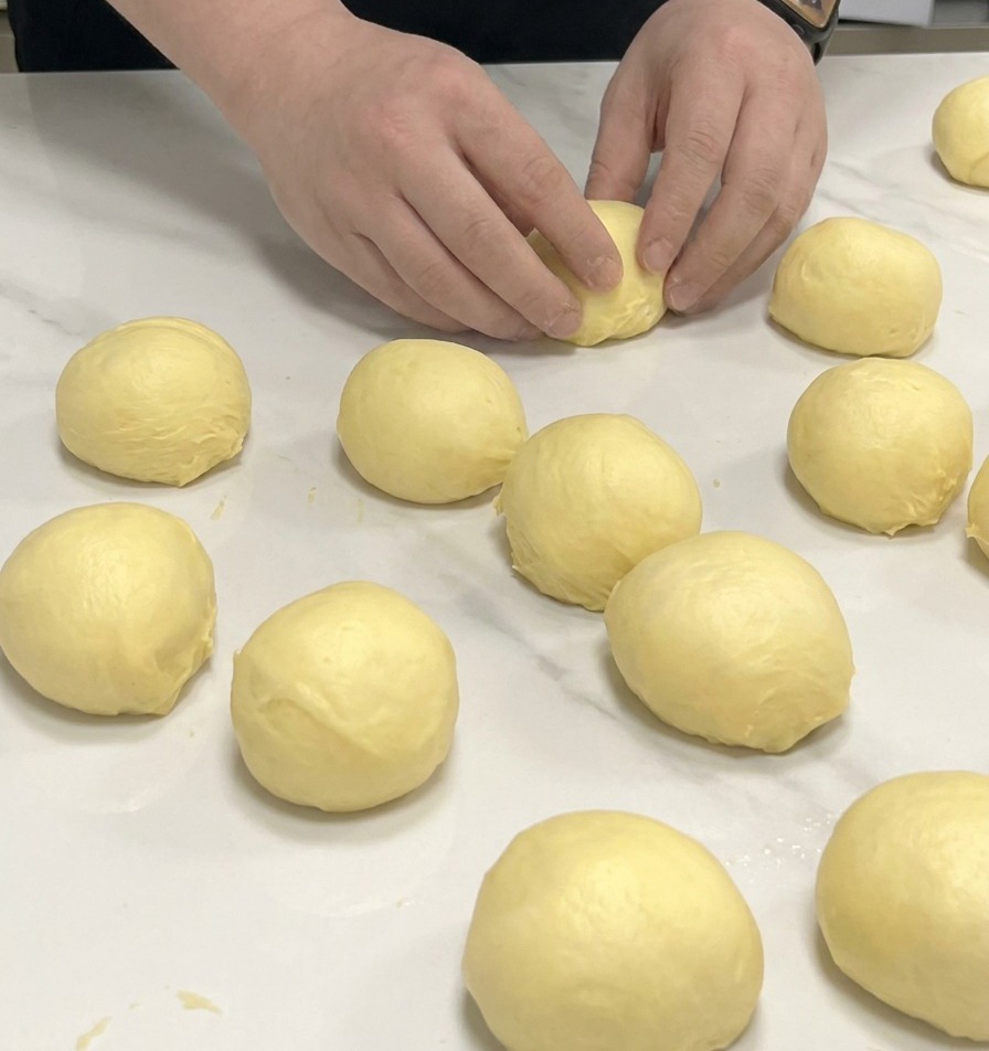 Hände formen kleine Portionen gelben Brioche-Teig auf einer Marmorplatte zu Kugeln.