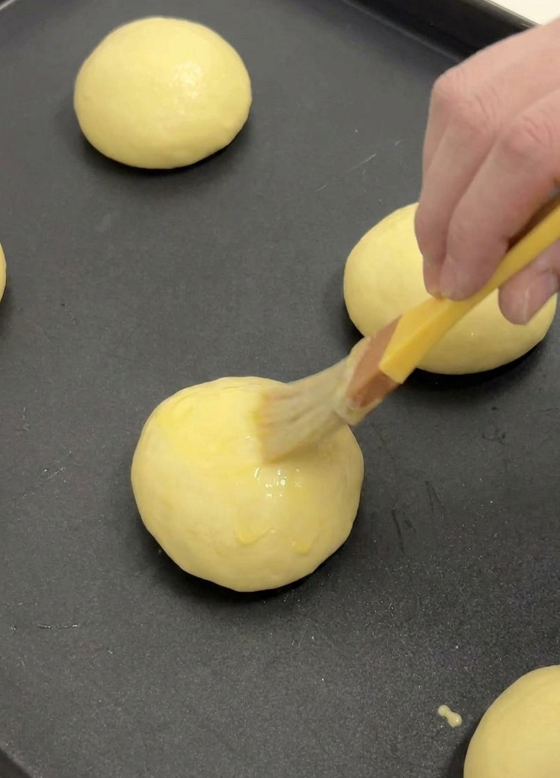 Ein Backpinsel trägt eine dünne Schicht Eigelb auf die Oberseite eines aufgegangenen Brioche-Buns auf.