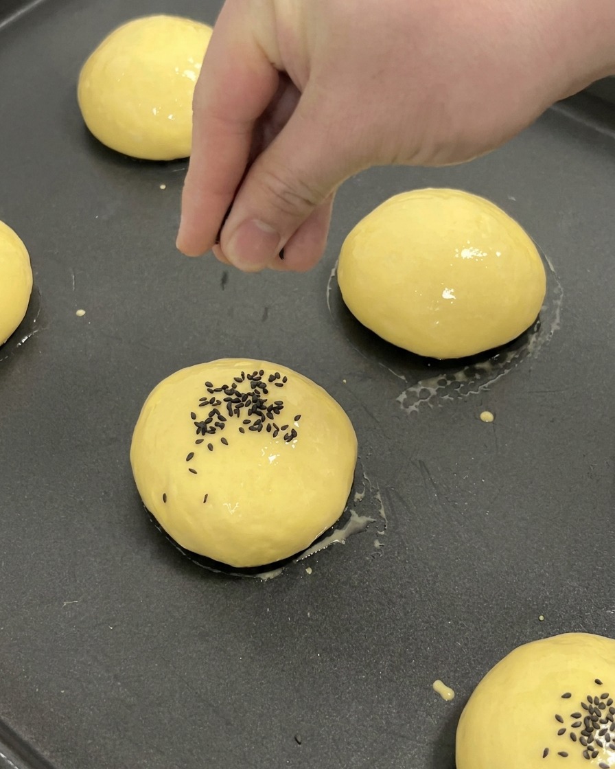 Eine Hand streut schwarzen Sesam in die Mitte eines mit Ei bestrichenen Brioche-Buns auf einem Backblech.