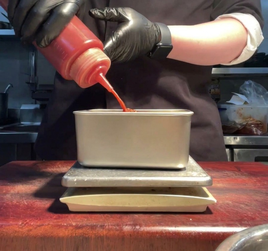 Hände in Handschuhen drücken hellrote Sriracha-Sauce aus einer Flasche in einen Metallbehälter.