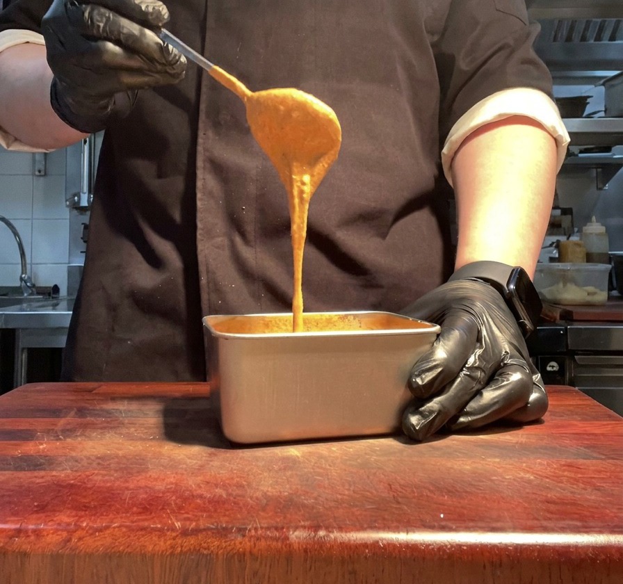 Eine Hand hebt einen Löffel glatter, orangefarbener Burgersauce aus einem Metallbehälter.