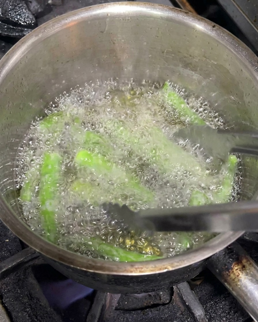 Metallzangen wenden grüne Chilis, während sie in einem Topf mit heißem Öl frittiert werden.