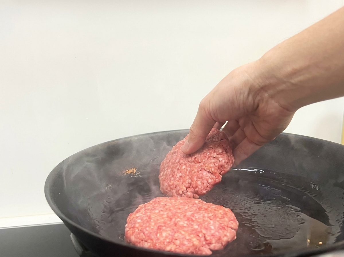 Eine Hand legt zwei rohe Rinder-Patties in eine heiße, leicht rauchende schwarze Bratpfanne.