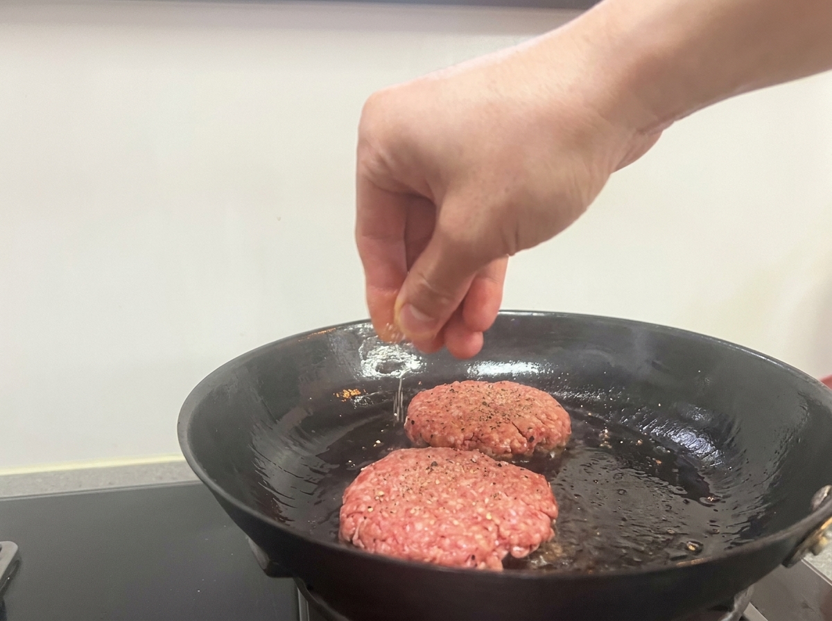 Eine Hand streut Salz und schwarzen Pfeffer über zwei bratende Patties in einer Pfanne.