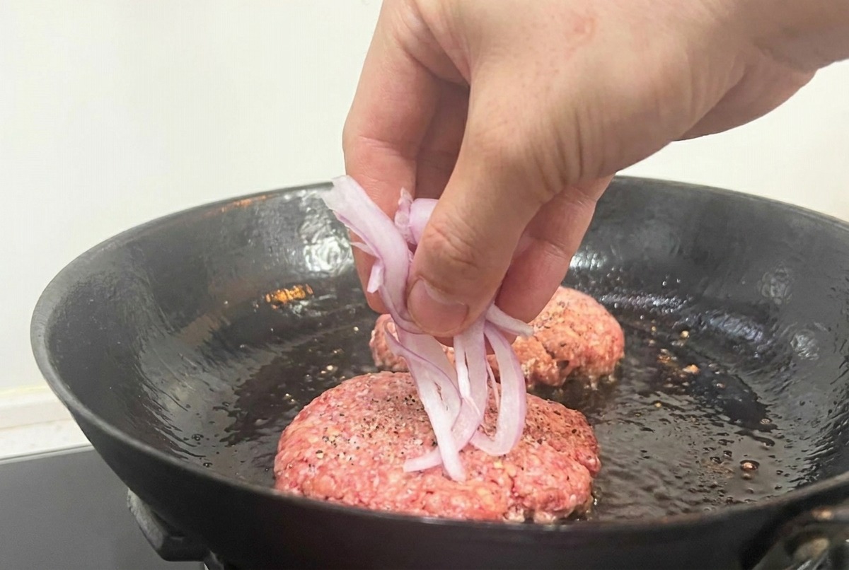 Eine Hand streut rote Zwiebelringe auf die rohen Fleisch-Patties in einer schwarzen Pfanne.