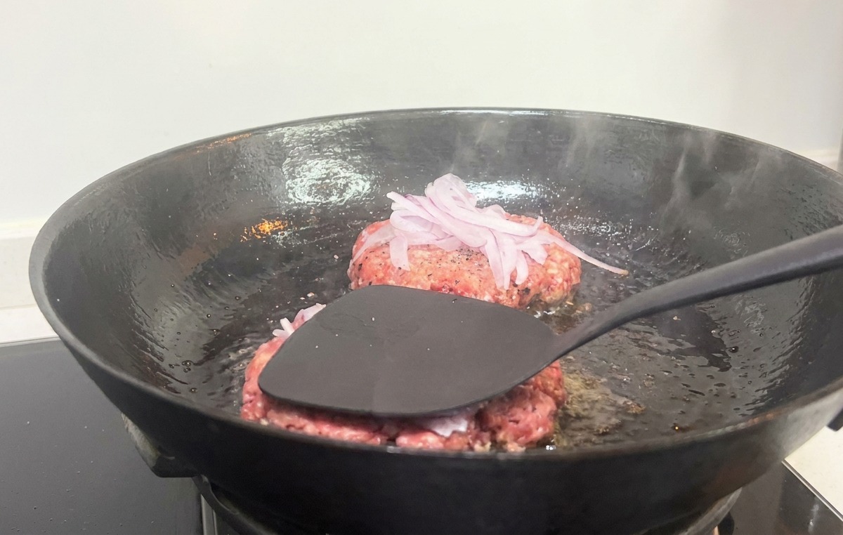 Ein schwarzer Pfannenwender drückt fest auf ein Burger-Patty mit roten Zwiebeln in einer heißen gusseisernen Pfanne.