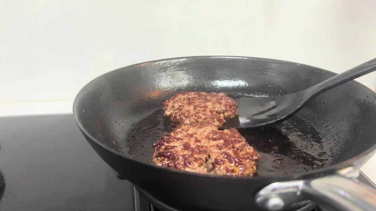 Ein schwarzer Pfannenwender wendet gebräunte Beef-Patties in einer Bratpfanne.