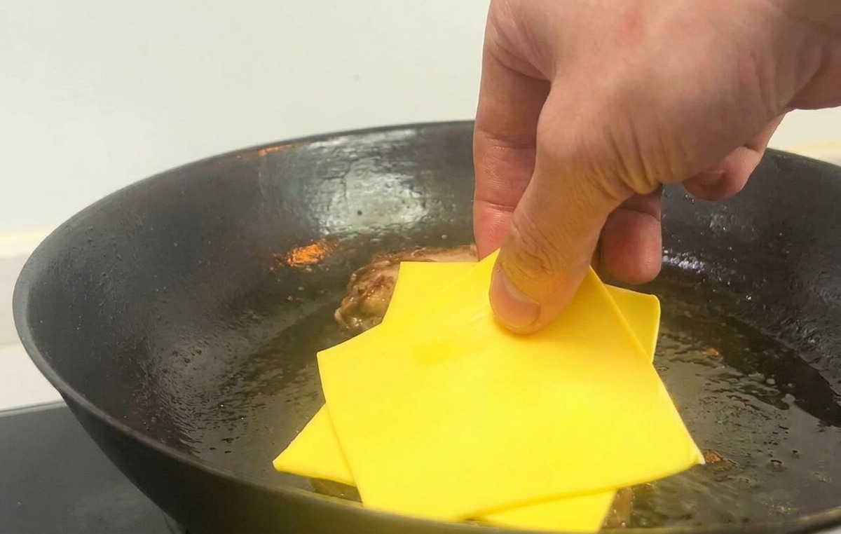 Eine Hand legt zwei quadratische Scheiben gelben Käse auf gebratene Burger-Patties in einer heißen Pfanne.