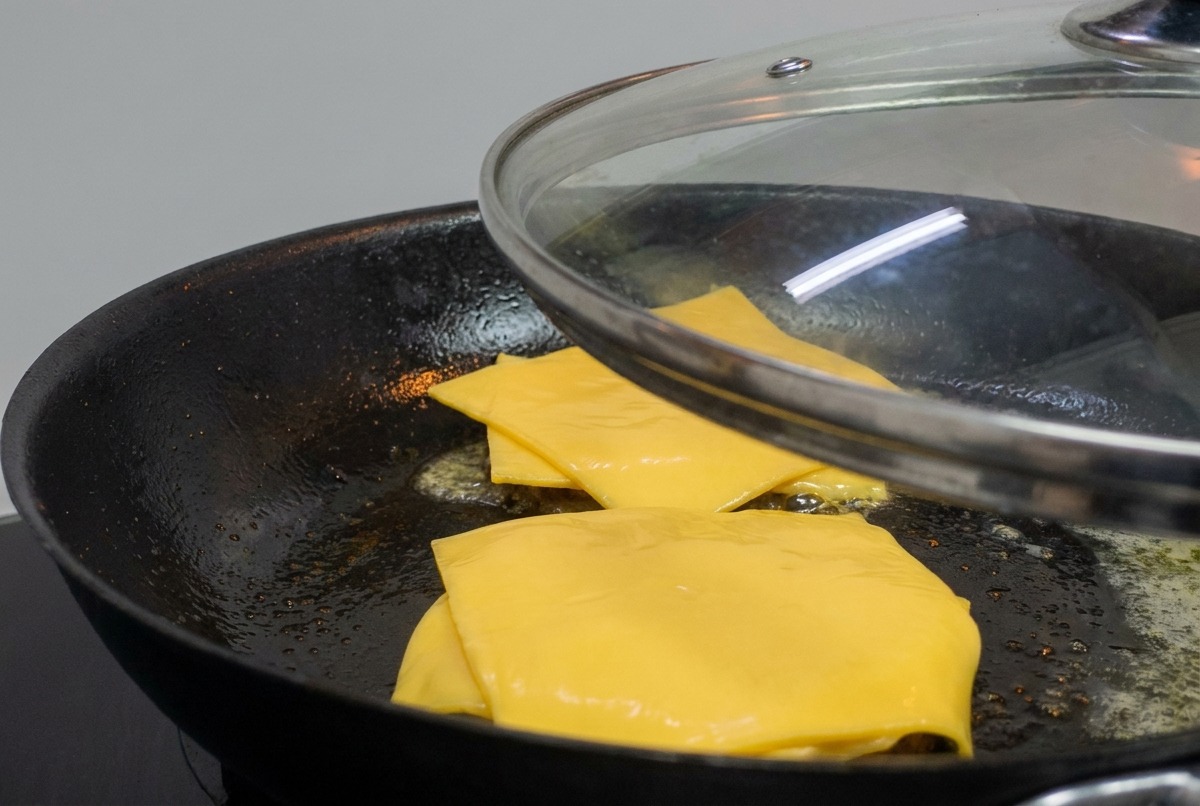 Ein Glasdeckel wird über eine Pfanne mit Burger-Patties und schmelzendem Käse gelegt.