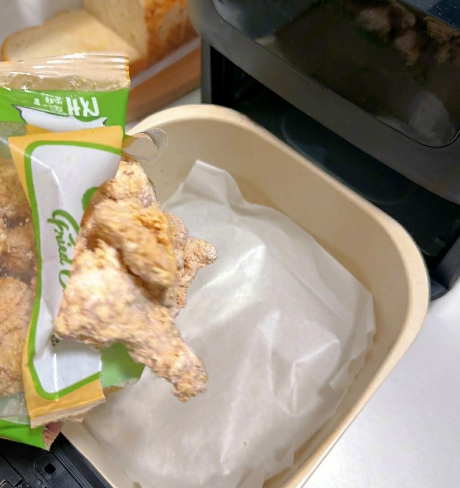 Gefrorene panierte Hähnchenteile werden aus einer Tüte in einen mit weißem Backpapier ausgelegten Heißluftfritteusenkorb geschüttet.