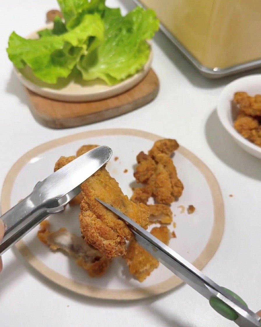 Nahaufnahme einer Küchenschere, die ein heißes Stück Fried Chicken schneidet, das mit einer Zange auf einem weißen Teller gehalten wird.