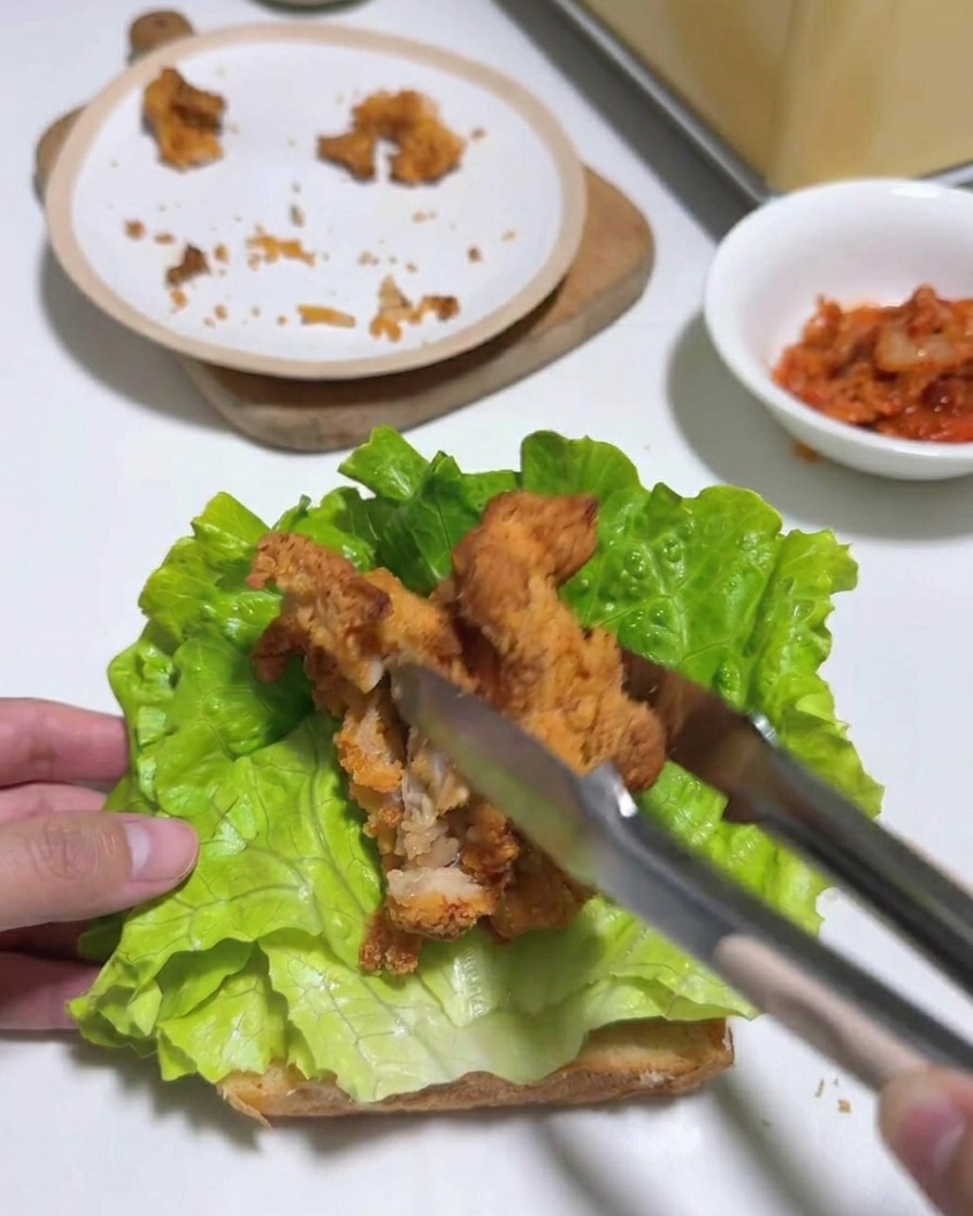 Eine Zange legt mehrere Streifen Fried Chicken auf eine Schicht Salat auf getoastetem Brot.
