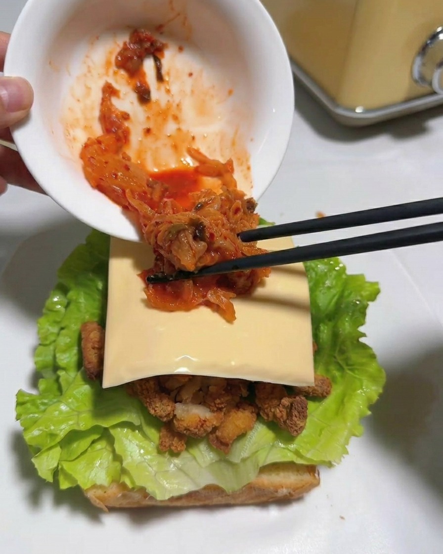 Stäbchen legen hellrotes, gehacktes Kimchi aus einer weißen Schüssel auf eine Käsescheibe in einem Sandwich.
