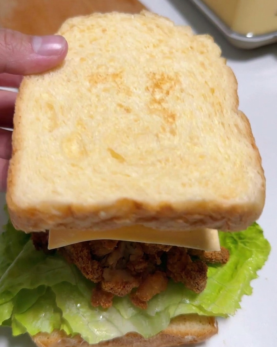 Eine Hand legt eine Scheibe getoastetes Brot auf ein fertiges Sandwich mit Salat, Hähnchen, Käse und Kimchi.