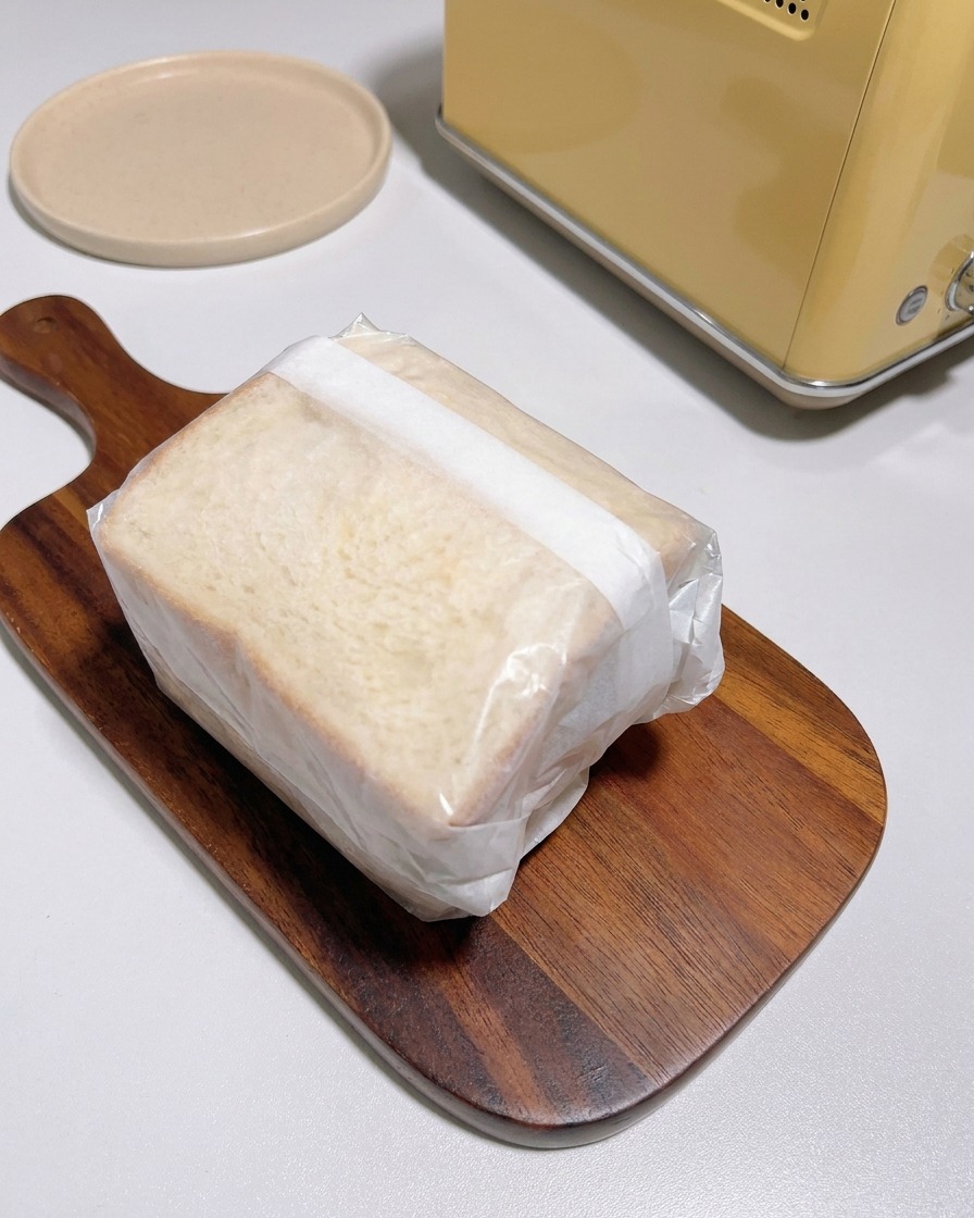 Ein dickes Sandwich, fest in weißes Pergamentpapier eingewickelt, liegt auf einem Holzbrett neben einem gelben Toaster.