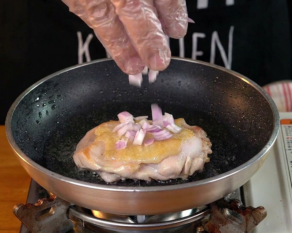 Eine Hand streut gewürfelte rote Zwiebeln in eine Pfanne zu einer bratenden Hähnchenkeule.