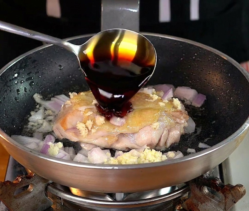 Ein Metalllöffel gießt dunkle Sojasauce über eine teilweise gegarte Hähnchenkeule in der Pfanne.