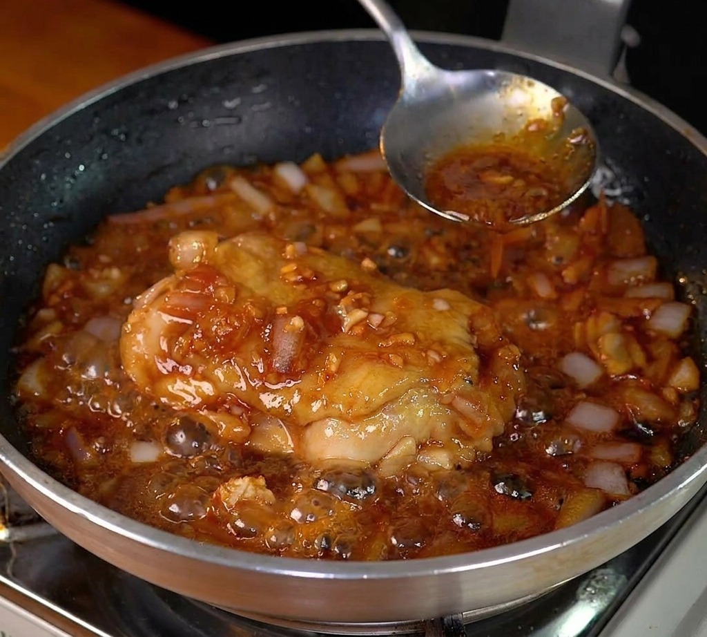 Hähnchenkeule köchelt in einer dunklen, blubbernden Sauce mit weichen Zwiebeln in einer Edelstahlpfanne.