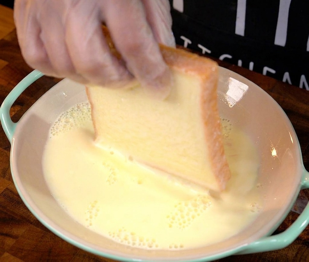 Eine Hand taucht eine dicke Scheibe Weißbrot in eine flache Schüssel mit Eiermilch.