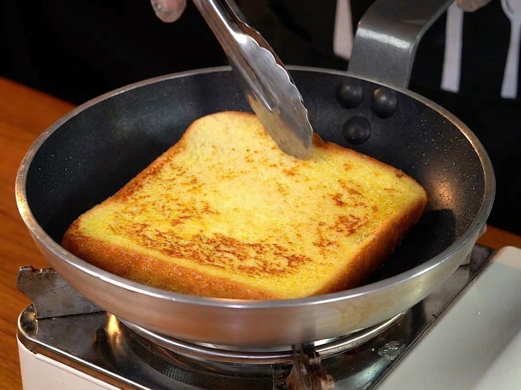 Eine Scheibe goldbrauner French Toast wird mit einer Metallzange in der Pfanne gewendet.