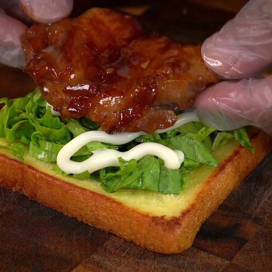 Behandschuhte Hände legen eine glänzende, karamellisierte Hähnchenkeule auf eine Sandwichbasis mit Salat und Mayonnaise.