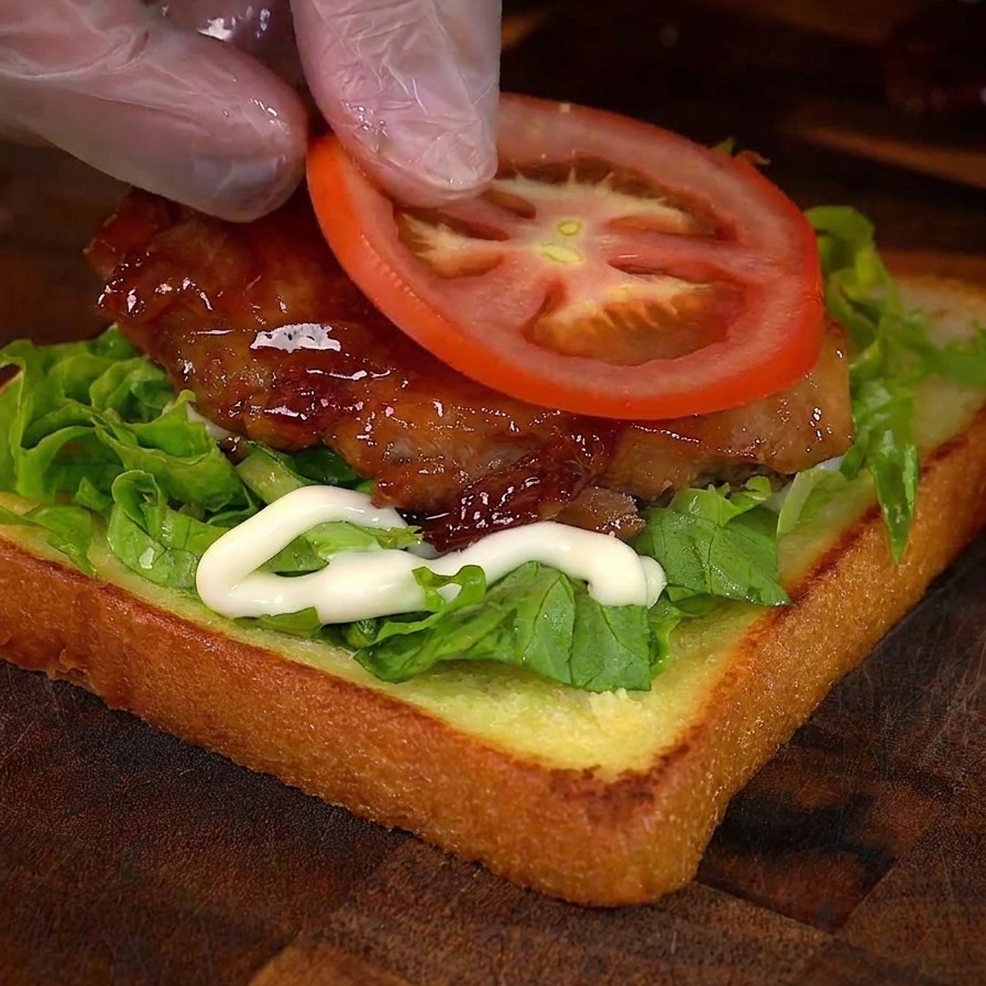 Eine Hand legt eine runde Scheibe frische Tomate auf ein Hähnchen-Sandwich.