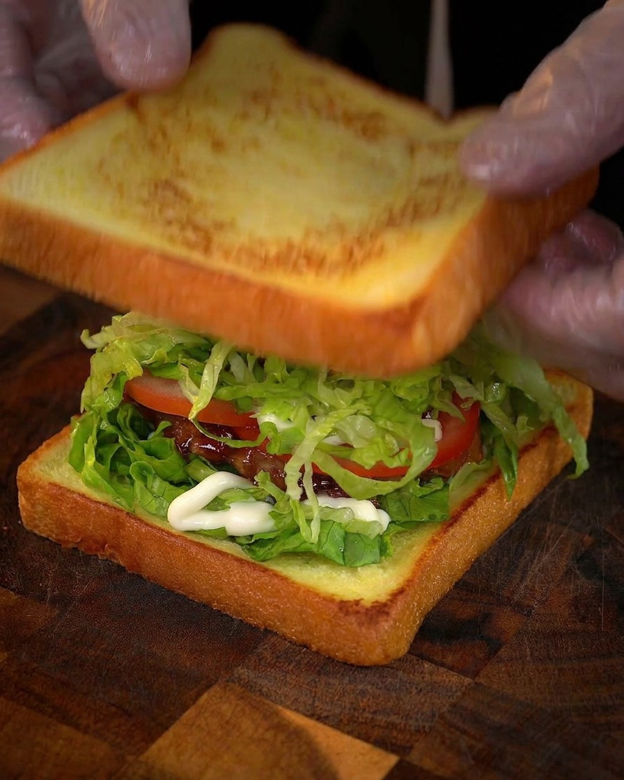 Behandschuhte Hände legen die zweite goldbraune Brotscheibe auf den Stapel aus Salat, Tomate und Hähnchen.