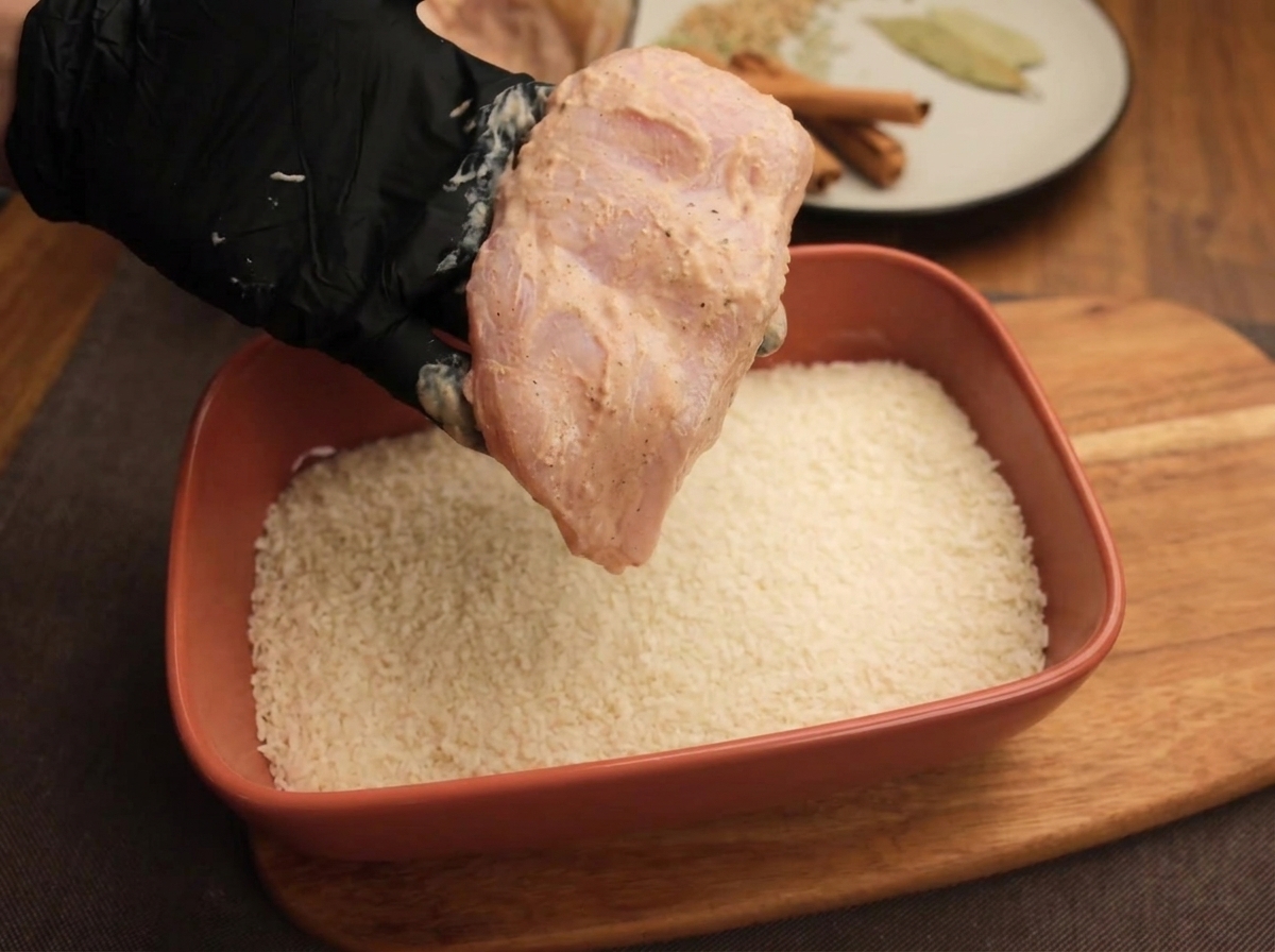 Eine Hand hält ein mariniertes Hähnchenschnitzel über eine Form mit weißen Semmelbröseln.